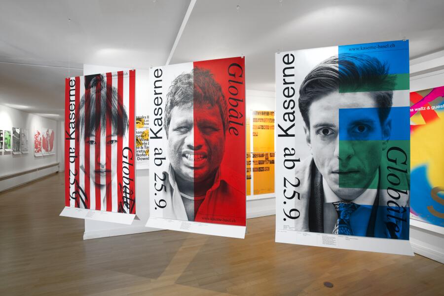 großformatige Plakate hängen diagonal in einem Ausstellungsraum. Die Plakate zeigen Gesichter, die mit farbigen Streifen und Flächen kombiniert wurden.