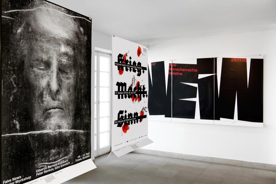 2 Plakate hängen von Schnüren von der Decke in einem Ausstellungsraum, ein weiteres hängt im Hintergrund an der Wand.
