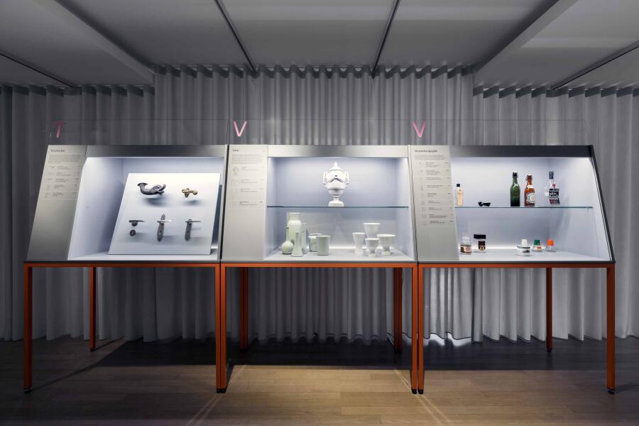 Vor einem hellgrauen Vorhang stehen 3 graue Vitrinen auf orangenen Beinen. Die Vitrinen sind von innen beleuchtet und zeigen Exponate aus den Sammlungen vom Focke Museum und dem Wilhelm Wagenfeld Haus.