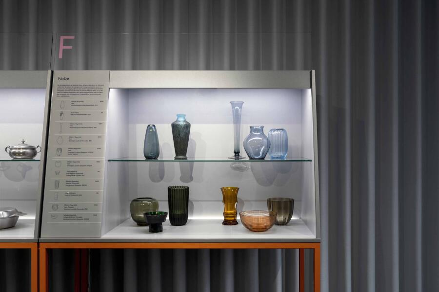 Vor einem hellgrauen Vorhang steht eine graue Vitrine auf orangenen Beinen. Die Vitrine ist von innen beleuchtet und zeigt  Vasen aus farbigem Glas.