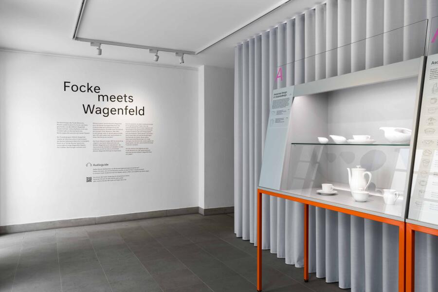 links zeigt das Foto einen schwarzen Wandtext vor einer weißen Wand, rechts sieht man eine graue Vitrine vor einem hellgrauen Vorhang.