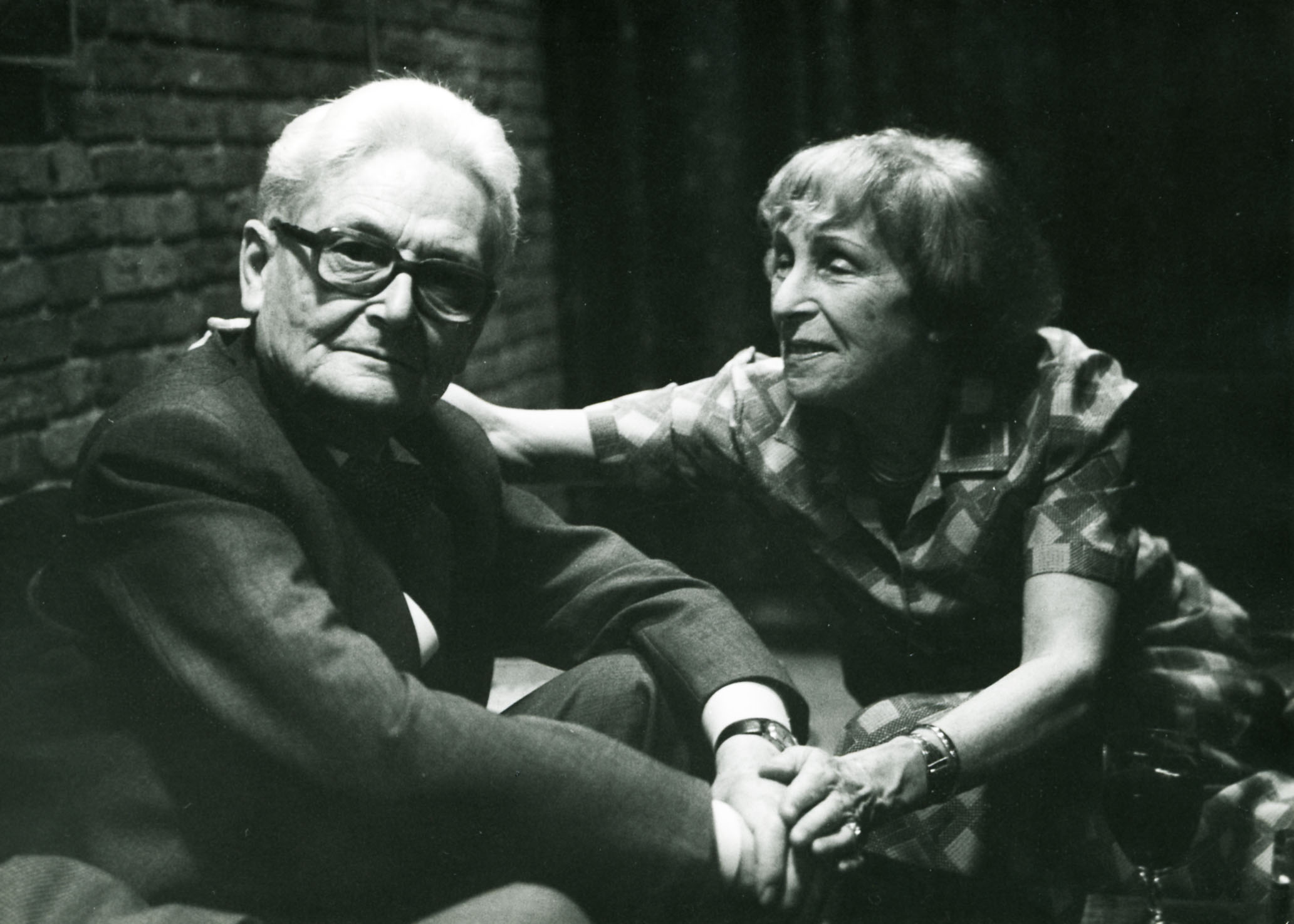 historisches schwarz-weiß Fot von 2 Personen, die auf einem niedrigen Sofa sitzen. Wilhelm Wagenfeld schaut in die Kamera, Margot Scharoun schaut auf Wagenfeld.