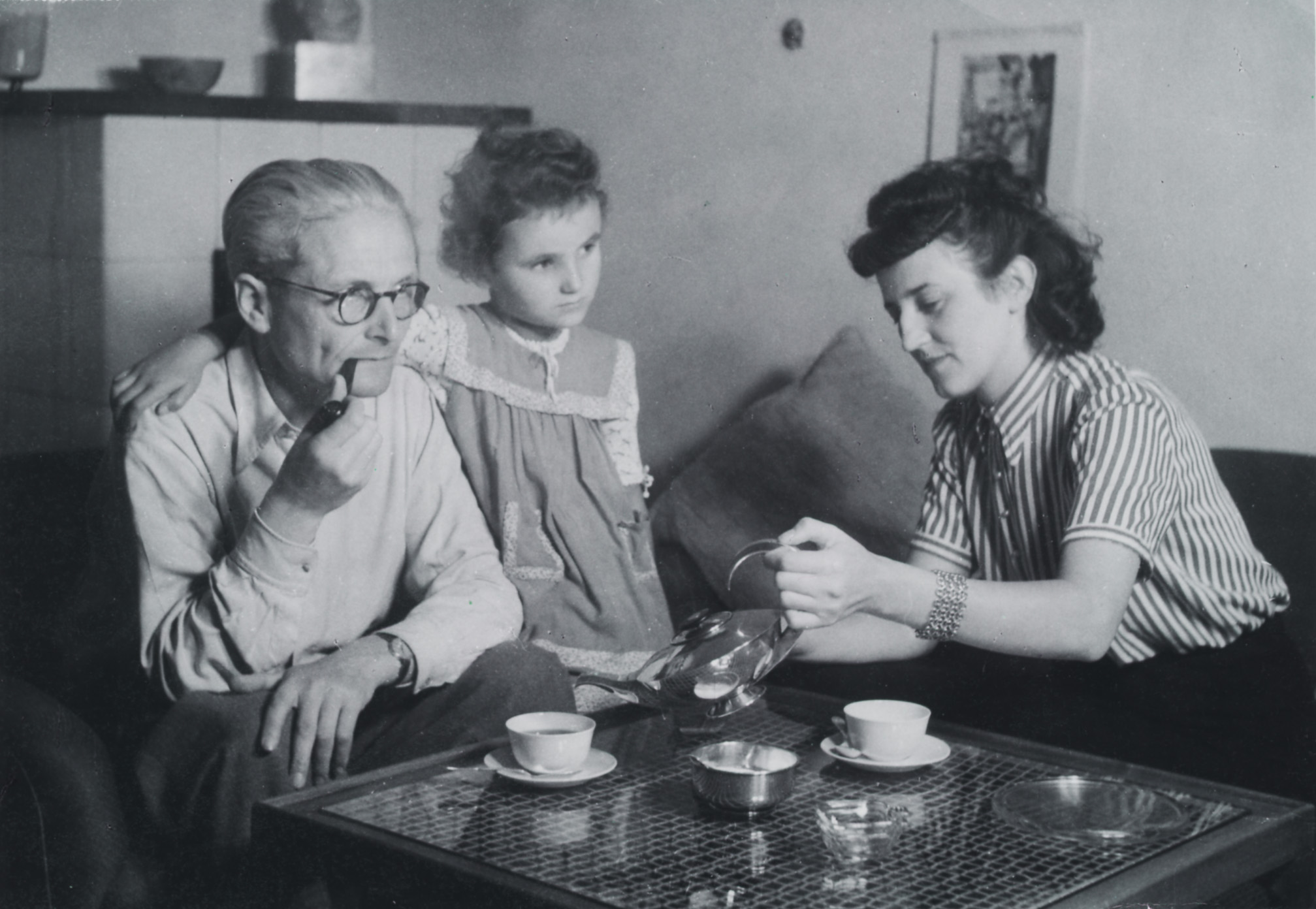 historisches schwarz-weiß-Foto von Vater, Mutter und Kind, die an einem niedrigen Kaffeetisch sitzen. Das Kind hat den rechten Arm um den Vater gelegt. 