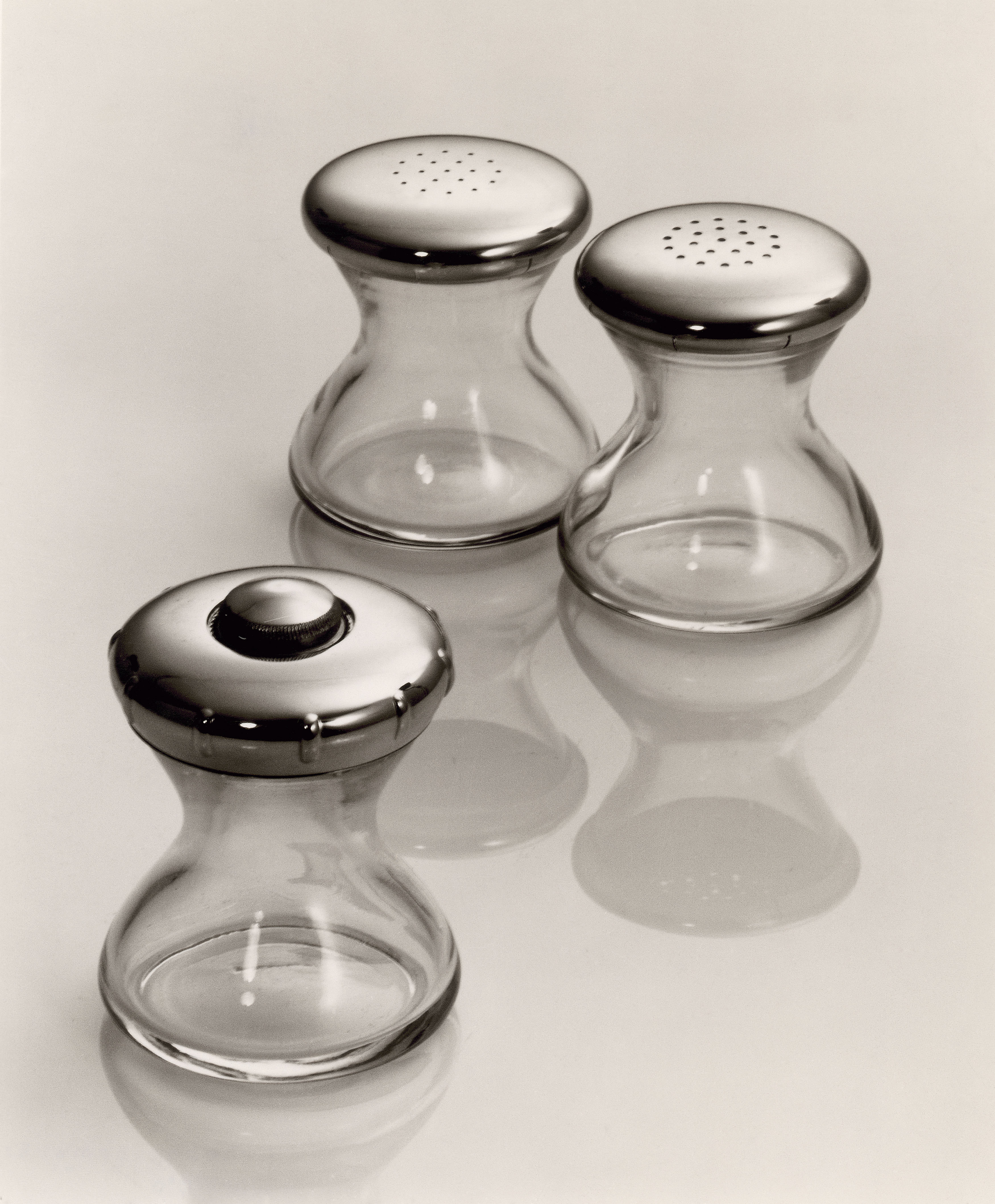 Salzstreuer, Pfefferstreuer und Pfeffermühle stehen auf einer spiegelnden Oberfläche. Die 3 Gefäße sind transparent, haben einen Deckel aus Edelstahl haben und teilen die gleiche elegant taillierte Form. 