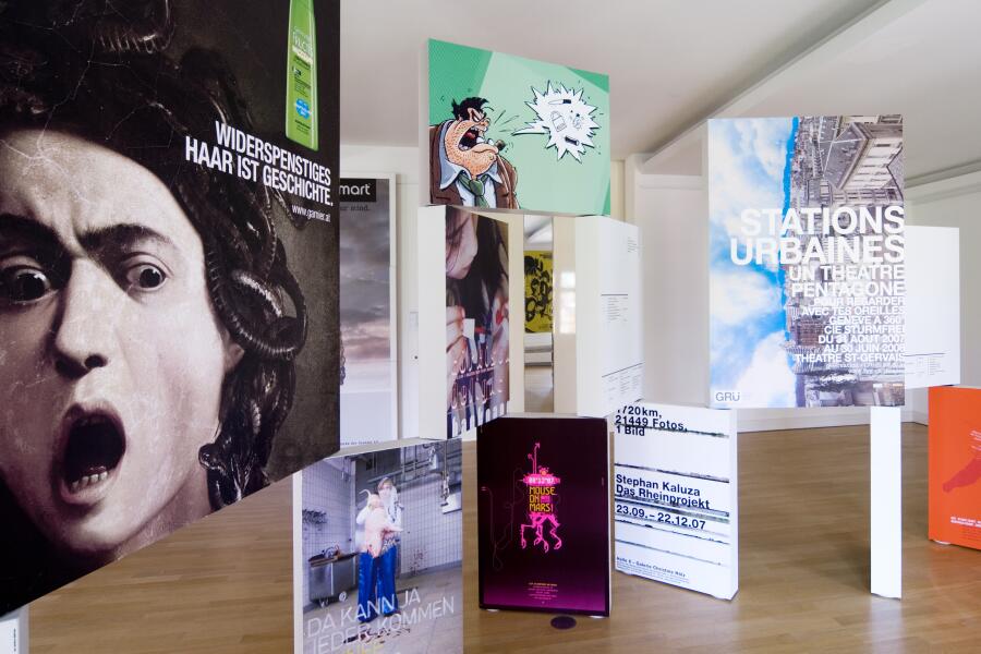 Bunte Plakate wurden auf schmalen, kastenförmigen Bildträgern aufgebracht und stehen nebeneinander und übereinander im Ausstellungraum.