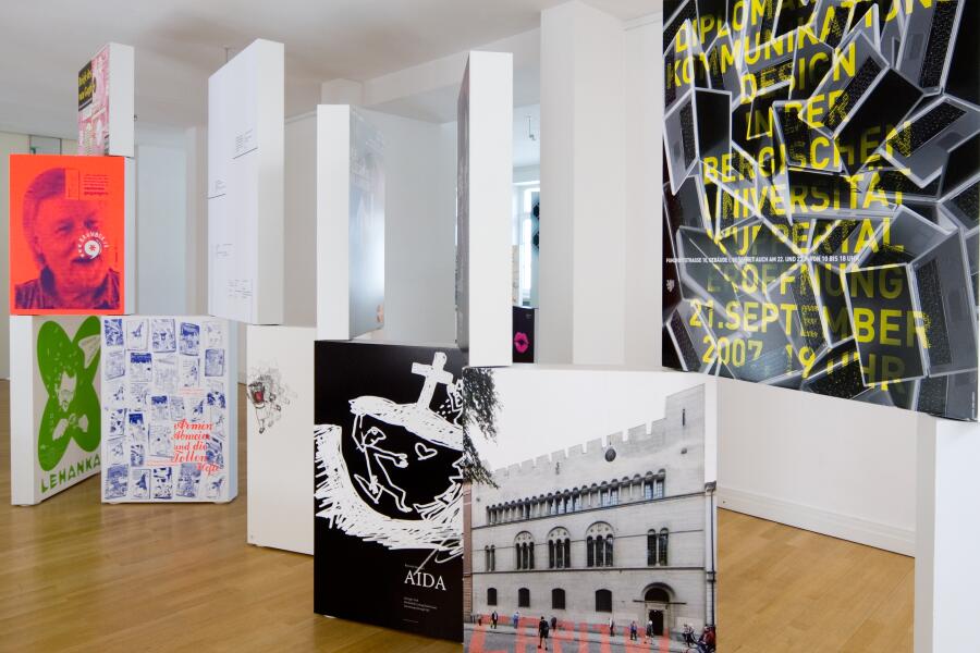 Bunte Plakate wurden auf schmalen, kastenförmigen Bildträgern aufgebracht und stehen nebeneinander und übereinander im Ausstellungraum.