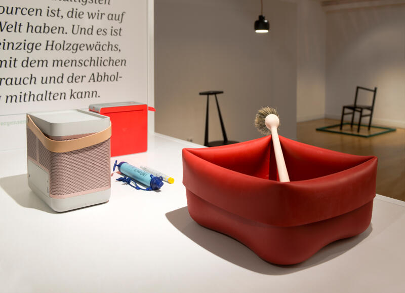 Auf einem Tisch stehen Exponate der Ausstellung, u.a. eine rote Ppülschüssel aus Gummi mit einer Bürste
