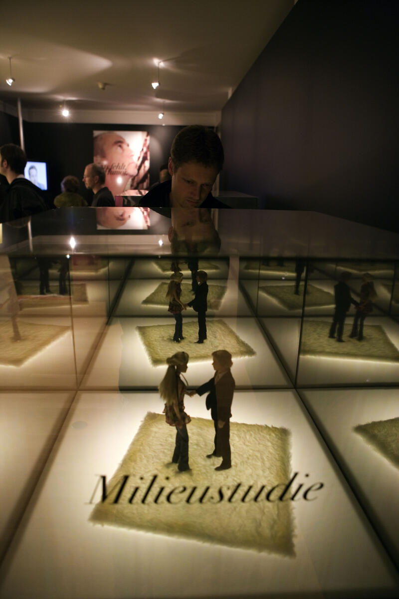 In einer Vitrine stehen zwei Figuren auf einem großen Blatt Papier. Auf der Vitrine steht in Schreibschrift das Wort Milieustudie.