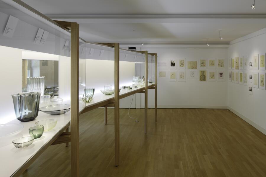 auf der linken Seite eine diagonal durch den Raum verlaufende Vitrine mit Wagenfeld-Objekten, auf der rechten Seite auf die Wand gehängte Entwurfszeichnungen.